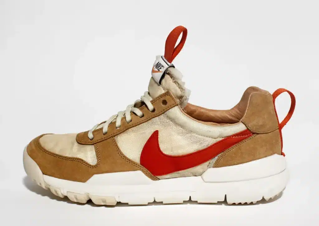 A stylish sneaker with a brown and beige color scheme, featuring a prominent red swoosh logo and red pull tabs on the tongue and heel, set against a white background. Tom Sachs Mars Yard 3.0