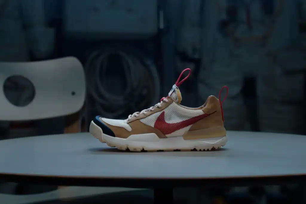 A sneaker with a beige and white body, red swoosh logo, and red pull tabs, displayed on a round table against a dark background. Tom Sachs Mars Yard 3.0