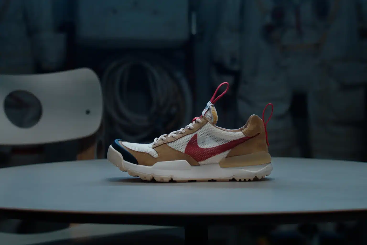 A sneaker with a beige and white body, red swoosh logo, and red pull tabs, displayed on a round table against a dark background. Tom Sachs Mars Yard 3.0