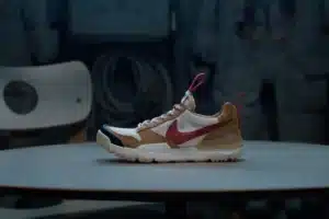 A sneaker with a beige and white body, red swoosh logo, and red pull tabs, displayed on a round table against a dark background. Tom Sachs Mars Yard 3.0