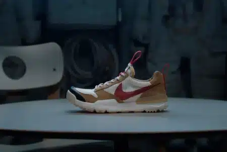 A sneaker with a beige and white body, red swoosh logo, and red pull tabs, displayed on a round table against a dark background. Tom Sachs Mars Yard 3.0