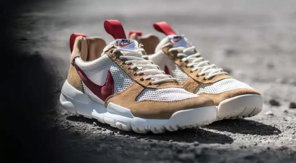 A pair of sneakers with a cream and beige color scheme, featuring red detailing and a prominent red swoosh logo on a mesh and suede upper, resting on a textured ground.