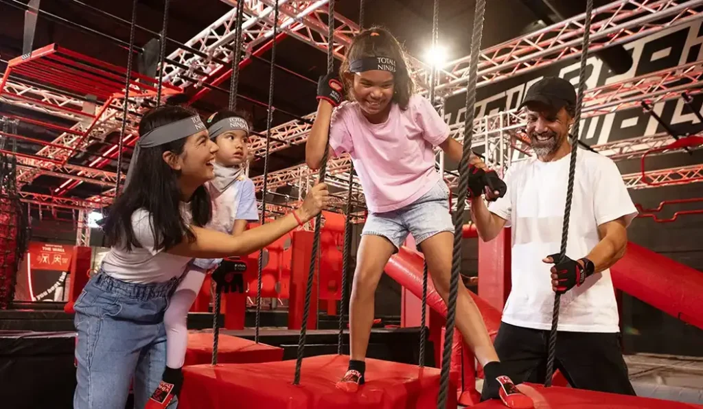 A family enjoying a ninja obstacle course indoors, with two children climbing a rope under the guidance of their parents. The course features red and black structures with ropes and platforms, and the family is wearing matching headbands and gloves.