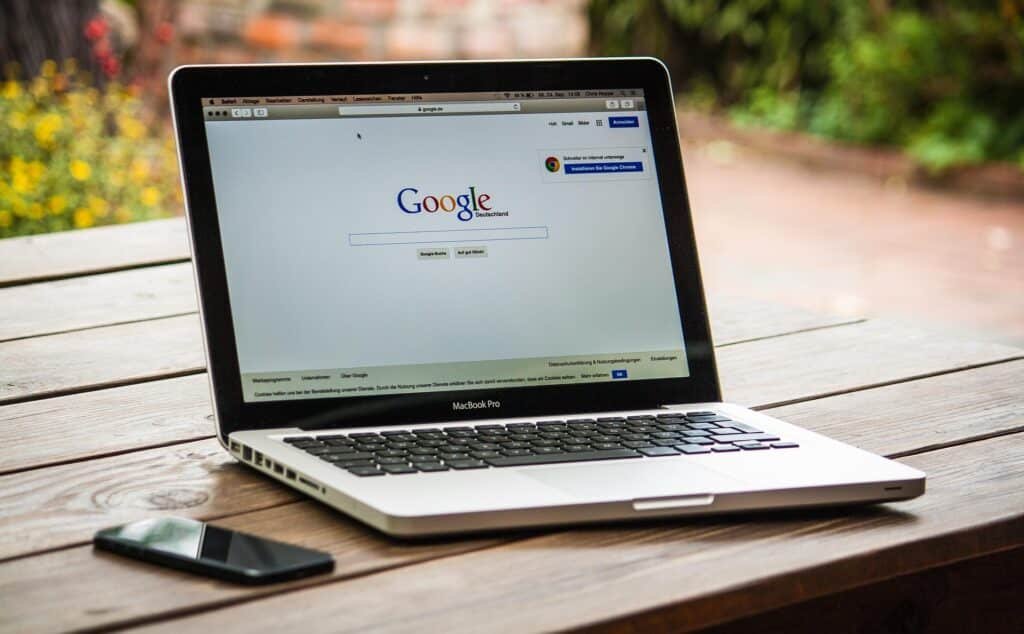 A MacBook Pro displaying the Google homepage in German is placed on a wooden outdoor table next to a smartphone.