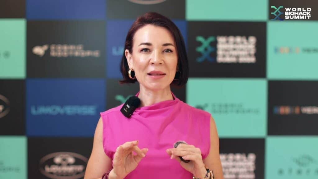 A woman in a bright pink sleeveless top speaking in front of a promotional backdrop for the World Biohack Summit, wearing a small microphone and large hoop earrings.