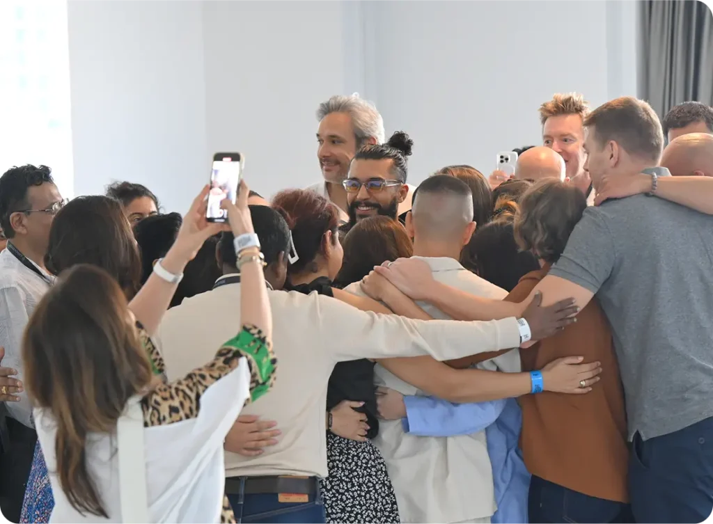 A group of people embracing in a communal hug, some taking photos with smartphones, in a well-lit indoor space. World Biohack Summit