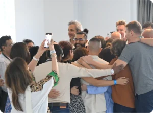 A group of people embracing in a communal hug, some taking photos with smartphones, in a well-lit indoor space. World Biohack Summit