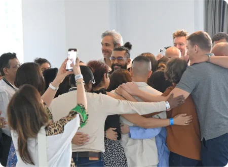 A group of people embracing in a communal hug, some taking photos with smartphones, in a well-lit indoor space. World Biohack Summit