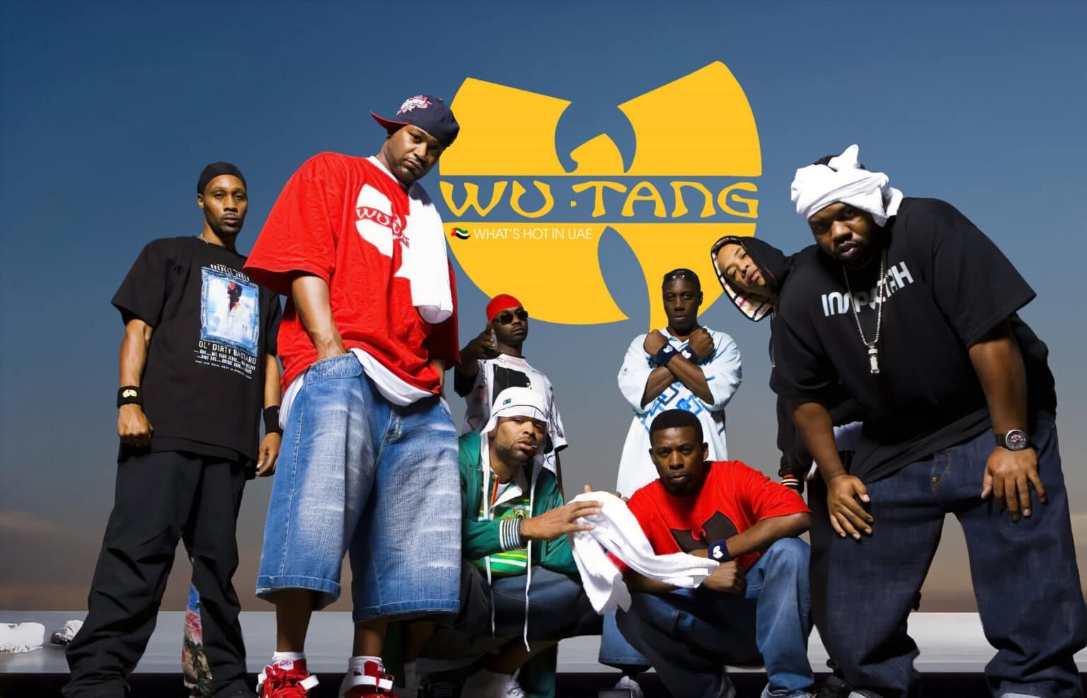 A group of eight men posing together, with a large yellow logo in the background that reads "WU-TANG," accompanied by "WHAT'S HOT IN UAE." They are dressed in casual streetwear, including jeans, t-shirts, and caps, with some draping towels over their shoulders. The background is a clear, blue sky.