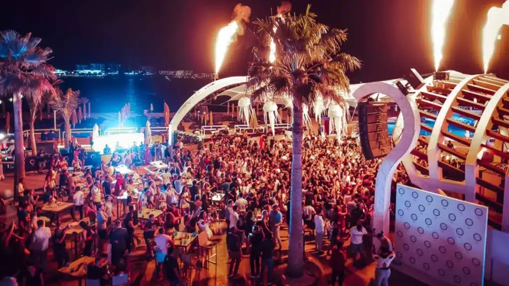 Nighttime beach club scene with a large crowd of people dancing under colorful lights and palm trees, featuring firework displays above and an outdoor DJ booth in the background.