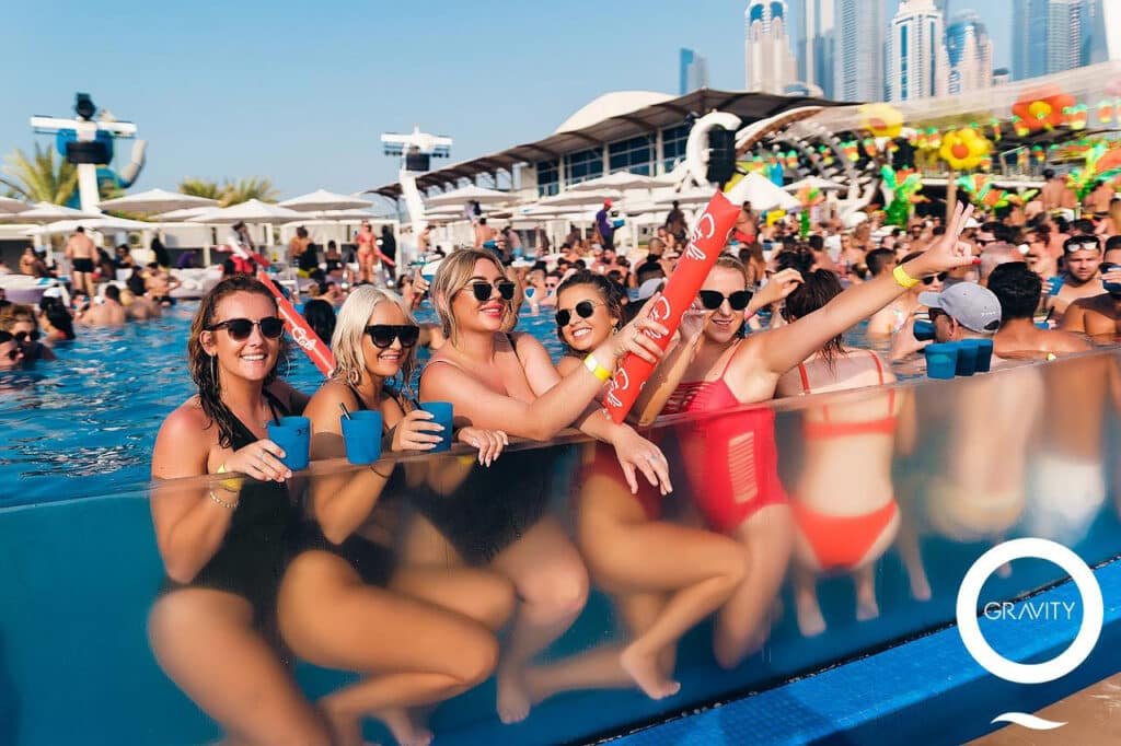 A group of women smiling and enjoying drinks at a pool party, surrounded by a lively crowd under a clear blue sky.