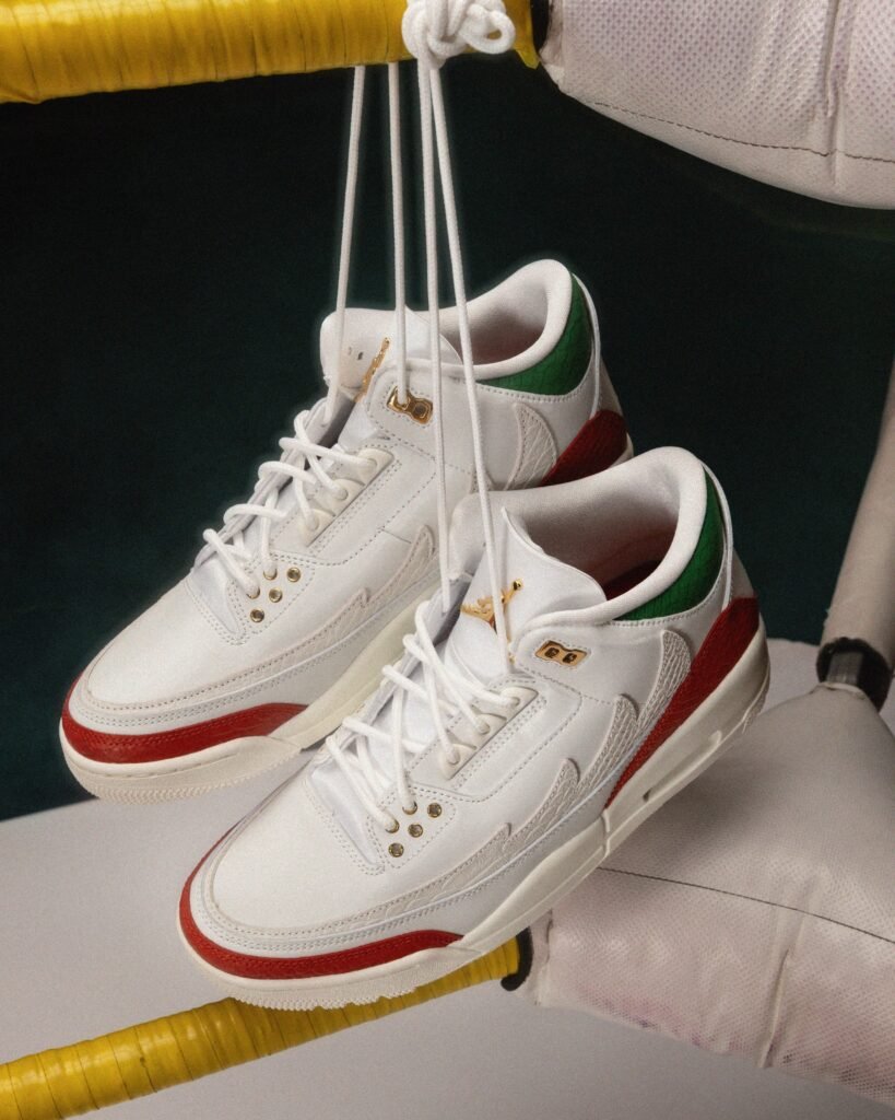 A pair of white sneakers with red, green, and gold accents hang by their laces over a bright yellow bar, with a cushioned corner in the background reminiscent of a boxing ring. Jordan El Vuelo Collection.