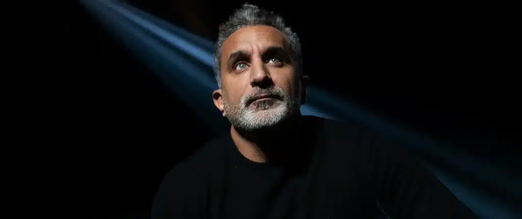 Bassem Youssef gazes upward under a focused beam of light against a dark background.