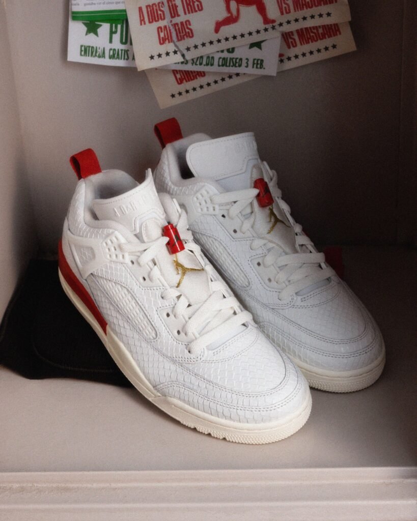 A pair of white sneakers with red accents and a gold logo, featuring a textured pattern, placed on a shelf with colorful posters partially visible in the background. Jordan El Vuelo Collection