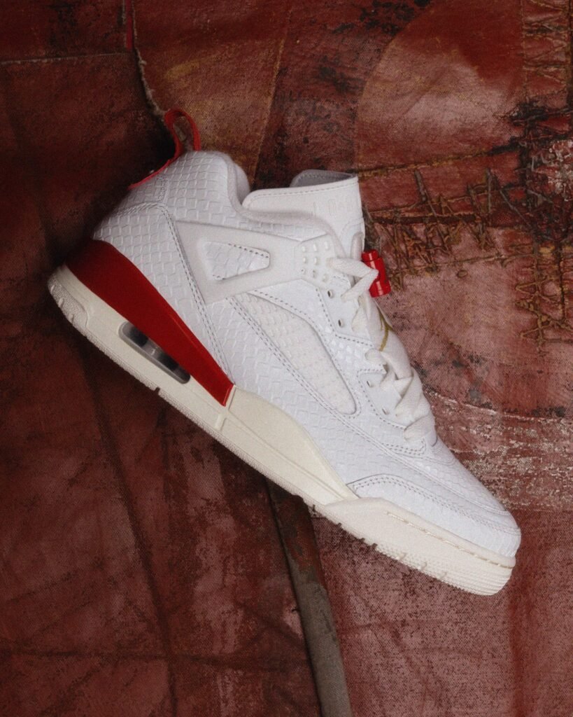 A white sneaker with textured detailing and a red heel accent, featuring an air cushion in the sole, placed against a textured reddish-brown background.