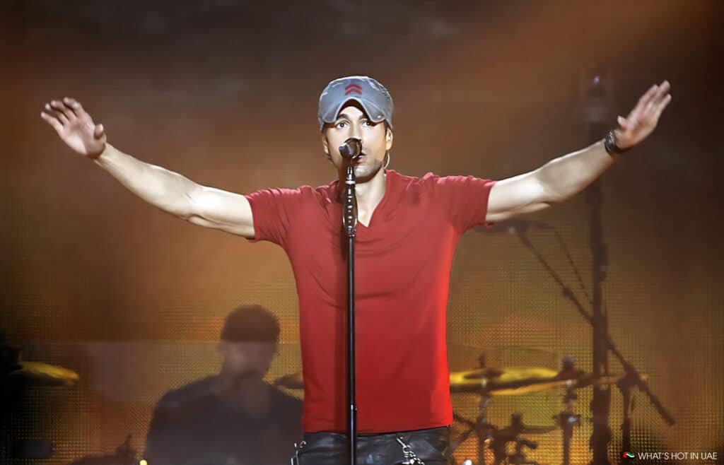 A man in a red shirt and gray cap performing on stage with his arms outstretched, standing in front of a microphone with a blurred drummer in the background.