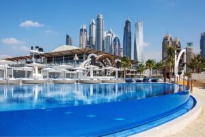 View of a blue swimming pool in Zero gravity Dubai, with white loungers and palm trees surrounding it. Modern skyscrapers are seen in the background under a clear blue sky.
