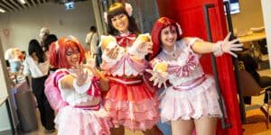 Three people in colorful anime-style costumes striking playful poses at a convention, with a background of other attendees and booths.