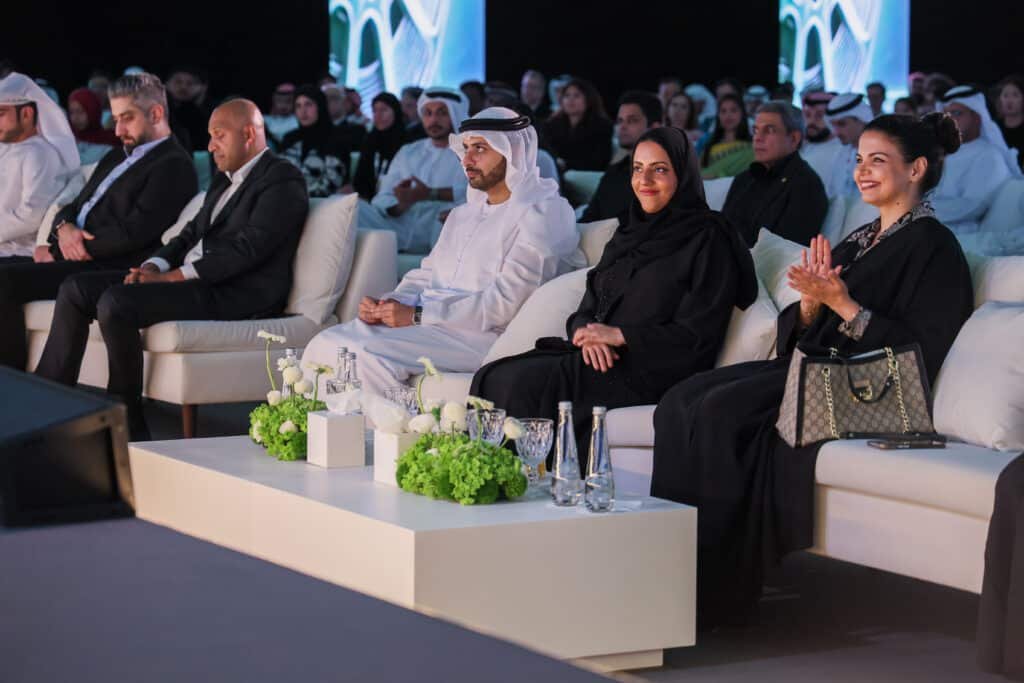 A group of people seated attentively in a conference setting, with some clapping and others listening, dressed in formal and traditional attire.