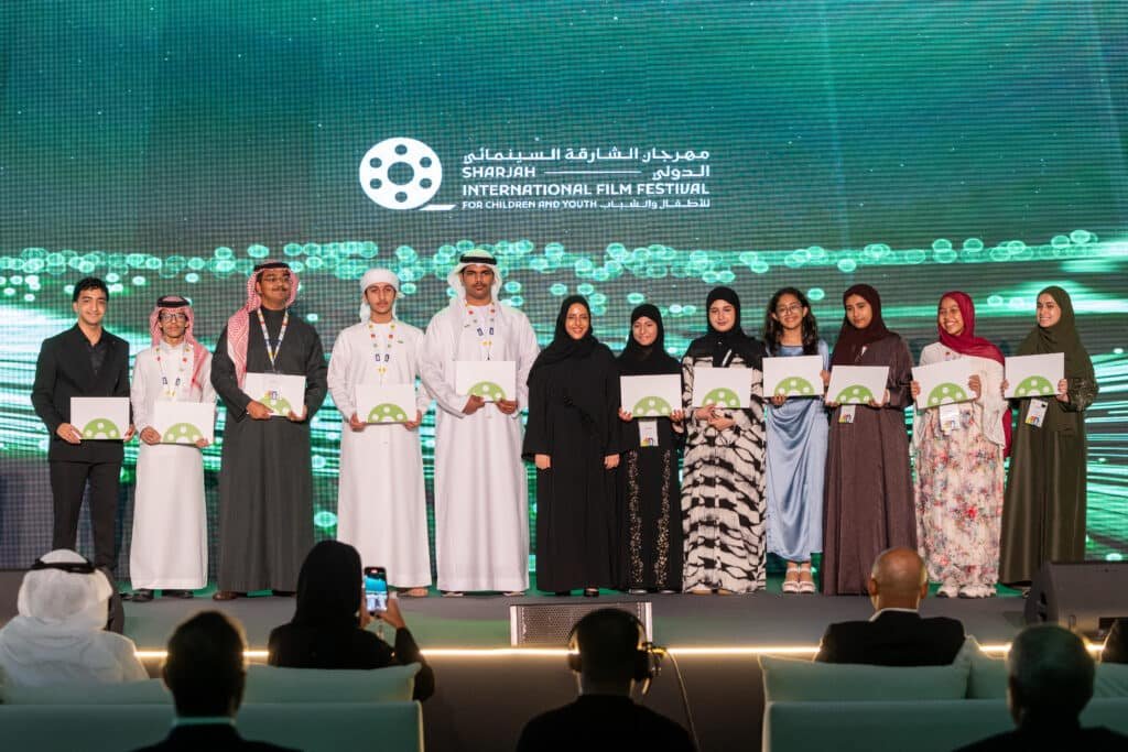 A group of people standing on stage holding certificates at the Sharjah International Film Festival for Children and Youth, with a green and white backdrop displaying the festival's name and logo.