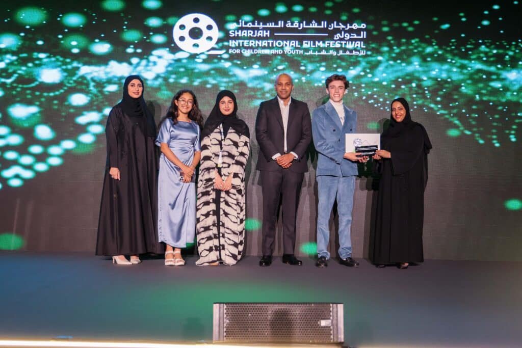 A group of six people standing on a stage at the Sharjah International Film Festival for Children and Youth. They are dressed formally with one person holding a certificate. The background features the festival's illuminated logo and design.