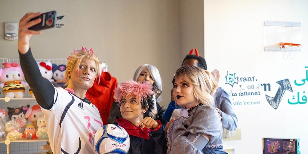 Geek Community Day Dubai: A group of people in a room taking a selfie. They are dressed in cosplay costumes, with colorful hairstyles and makeup, standing in front of shelves filled with plush toys. A small basketball hoop is mounted on the wall beside them.