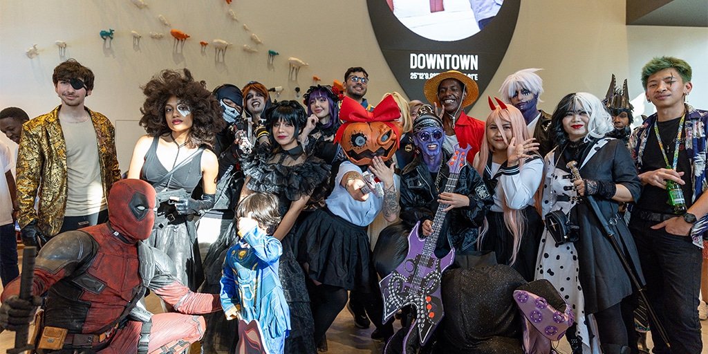 Geek Community Day Dubai: A group of people in diverse and colorful costumes posing at a cosplay event. The costumes include a mix of superheroes, anime characters, and unique creative outfits. The setting appears to be a convention hall with decorative elements on the wall and a digital display in the background.