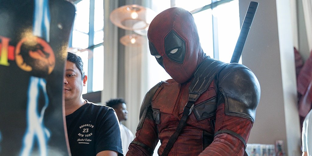 Geek Community Day Dubai: Person dressed as the superhero character Deadpool, wearing a red and black costume with a mask, standing inside a building with large windows and overhead lights.