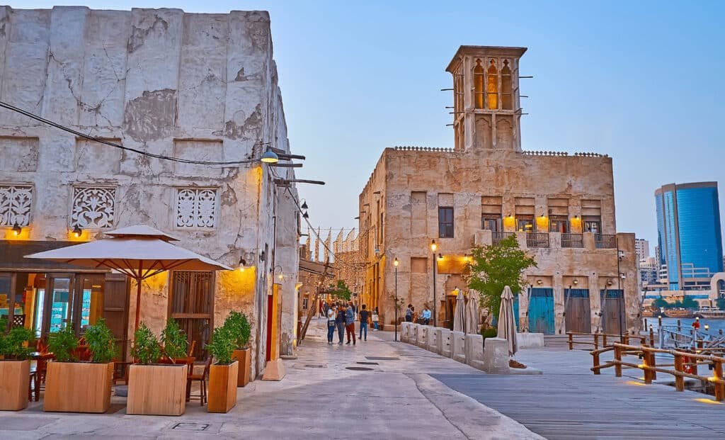 Historic district with traditional stone buildings, outdoor seating with umbrellas, and people walking along a cobblestone path under evening lighting.