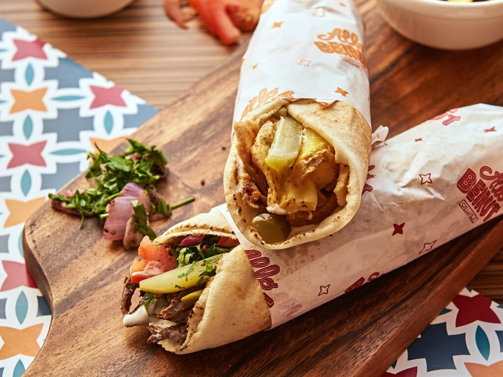 Late Night Food in Dubai: Two wrapped shawarma sandwiches on a wooden board, filled with vegetables and meat, with chopped parsley and onion pieces nearby.