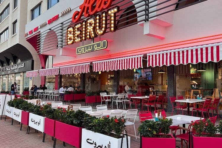 Outdoor seating area of a restaurant named 'Al Beirut' with red and white signage and striped awning, featuring red and white tables and chairs, surrounded by planters filled with greenery.