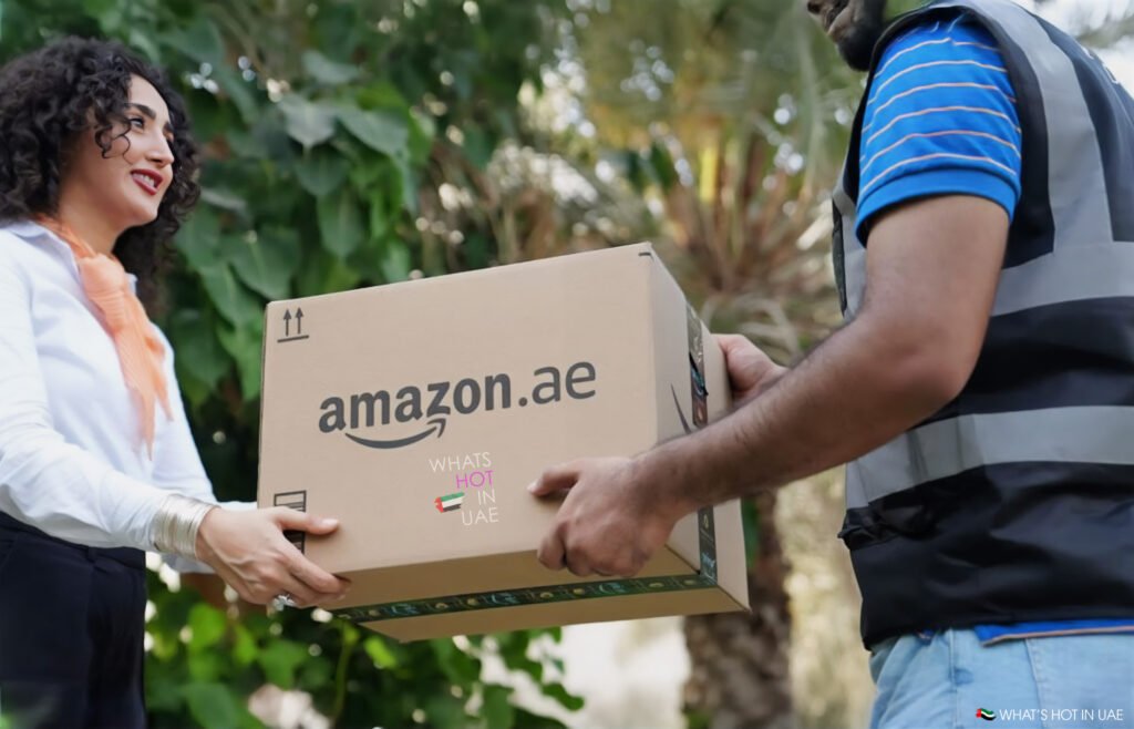 A woman receiving a package labeled 'amazon.ae' from a delivery person outdoors, surrounded by greenery.