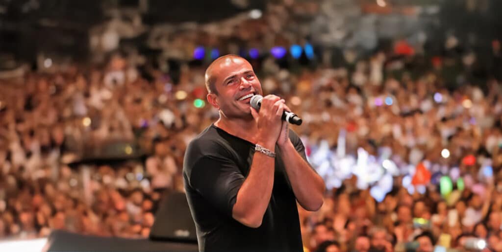 Amr Diab on stage holding a microphone, smiling while looking up, with a large, blurred audience in the background.