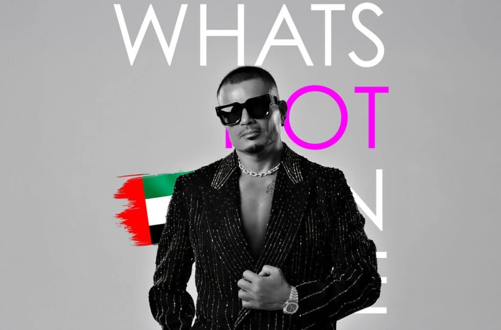 Amr Diab wearing a sparkling black suit and sunglasses, set in front of the United Arab Emirates flag, with the words "WHAT'S HOT" in bold letters.