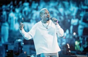 Amr Diab singing on stage with a microphone, wearing a white sweater and a necklace, against a blue-lit background.