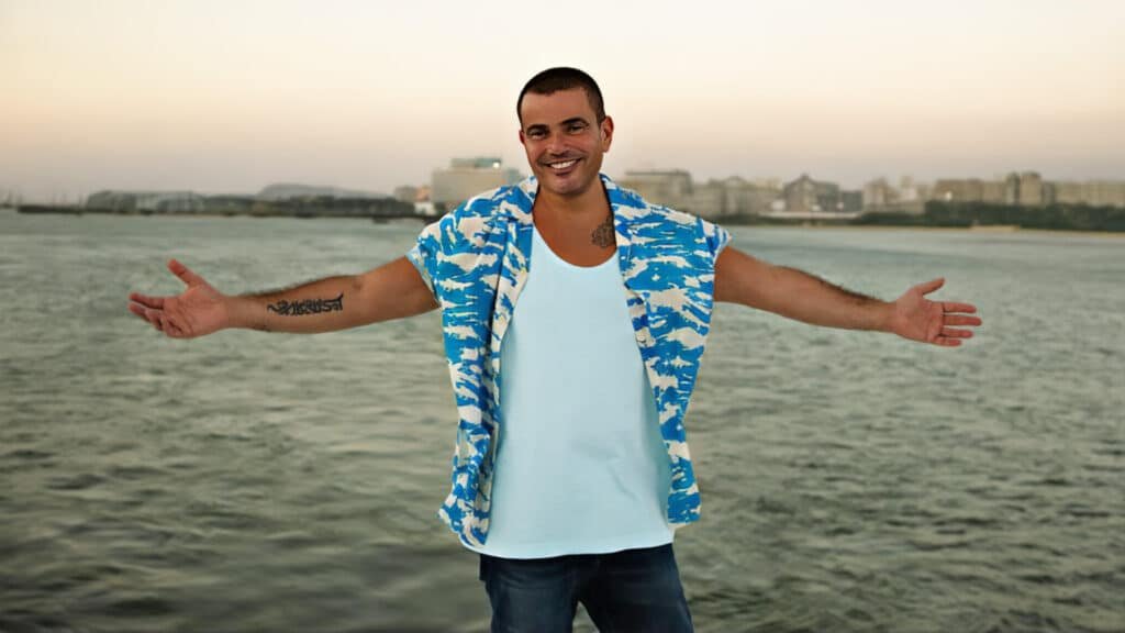 Amr Diab stands with arms wide open, wearing a blue and white patterned shirt over a light blue tank top, against a backdrop of water and distant city buildings.