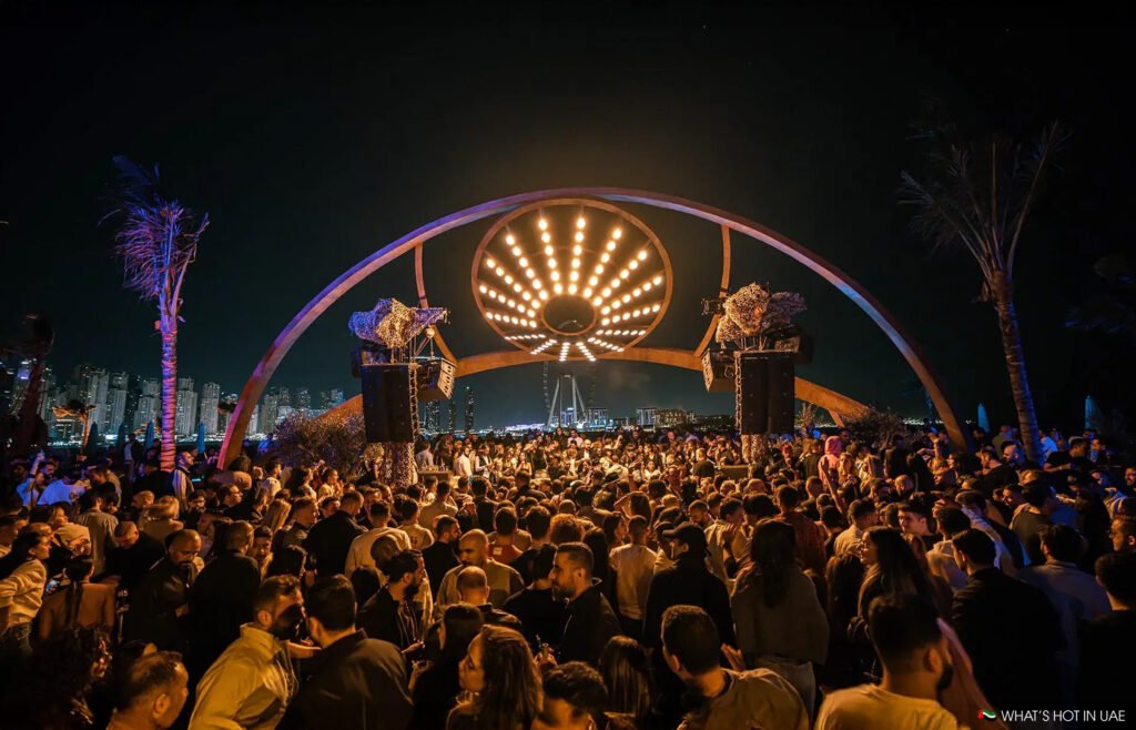 A large crowd gathered under an illuminated arch displaying a starburst pattern of lights, set against a cityscape backdrop at night.