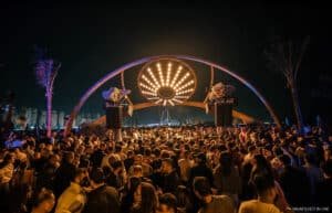 A large crowd gathered under an illuminated arch displaying a starburst pattern of lights, set against a cityscape backdrop at night.
