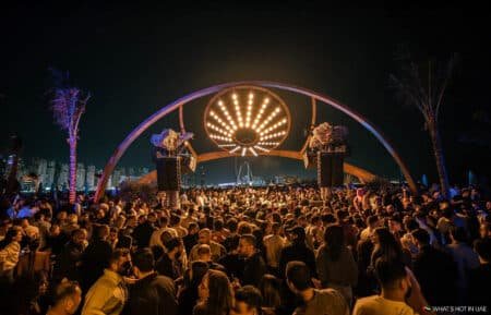 A large crowd gathered under an illuminated arch displaying a starburst pattern of lights, set against a cityscape backdrop at night.