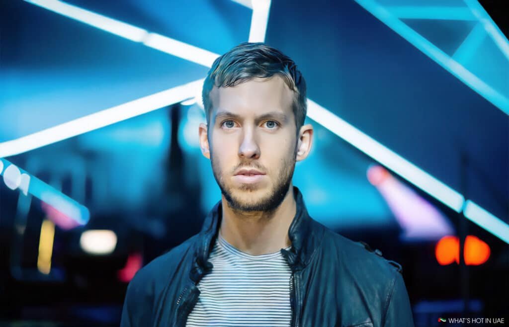Calvin Harris, wearing a striped shirt and a leather jacket, standing against a background of vibrant blue and white lights arranged in geometric patterns.