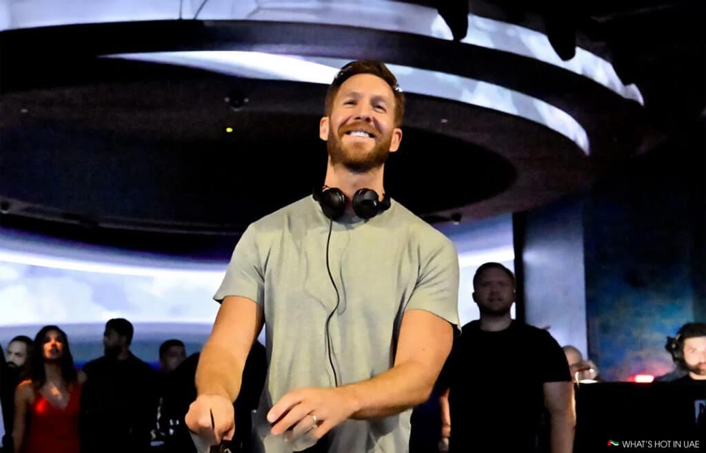 Calvin Harris is performing on stage with headphones around his neck, smiling, at a nightclub with a futuristic ceiling design and an audience gathered behind him.