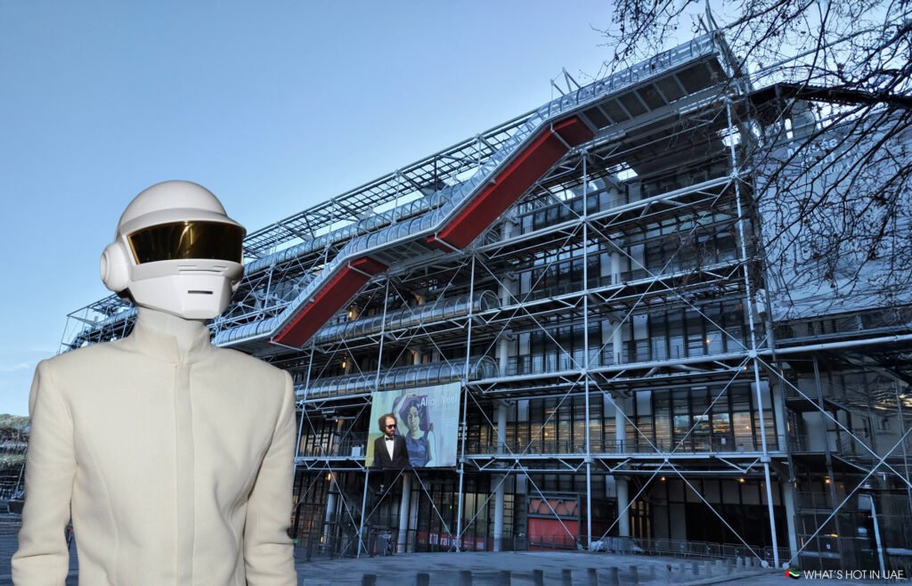 A person wearing a white helmet and jacket standing in front of the Centre Pompidou in Paris, a building known for its high-tech architecture with visible pipes and steel framework.