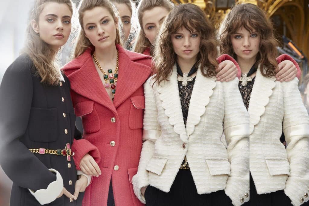 Group of models wearing stylish coats, including a pink textured coat and a black coat with gold buttons and colorful belt, posing together with arms around each other.