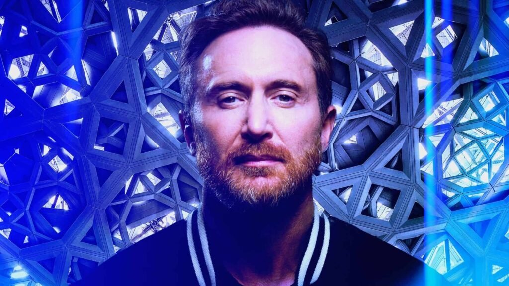 A man with a beard and short hair stands in front of a complex geometric background filled with interlocking triangular shapes and a blue lighting effect. David Guetta Ushuaia Dubai