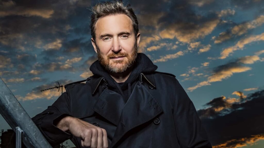 A man with short hair and a beard wearing a dark coat and hoodie stands outdoors under a dramatic sky with scattered clouds at sunset.