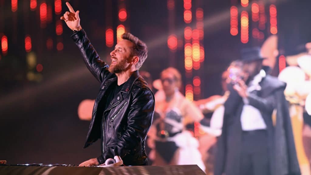A DJ in a black leather jacket is performing with one hand raised, surrounded by dancers in costumes on stage with vibrant red lighting in the background.