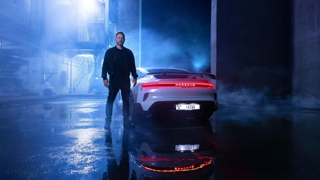 A man dressed in black stands beside a white Porsche Taycan in an industrial setting at night, surrounded by blue fog with wet pavement reflecting the car's rear lights.