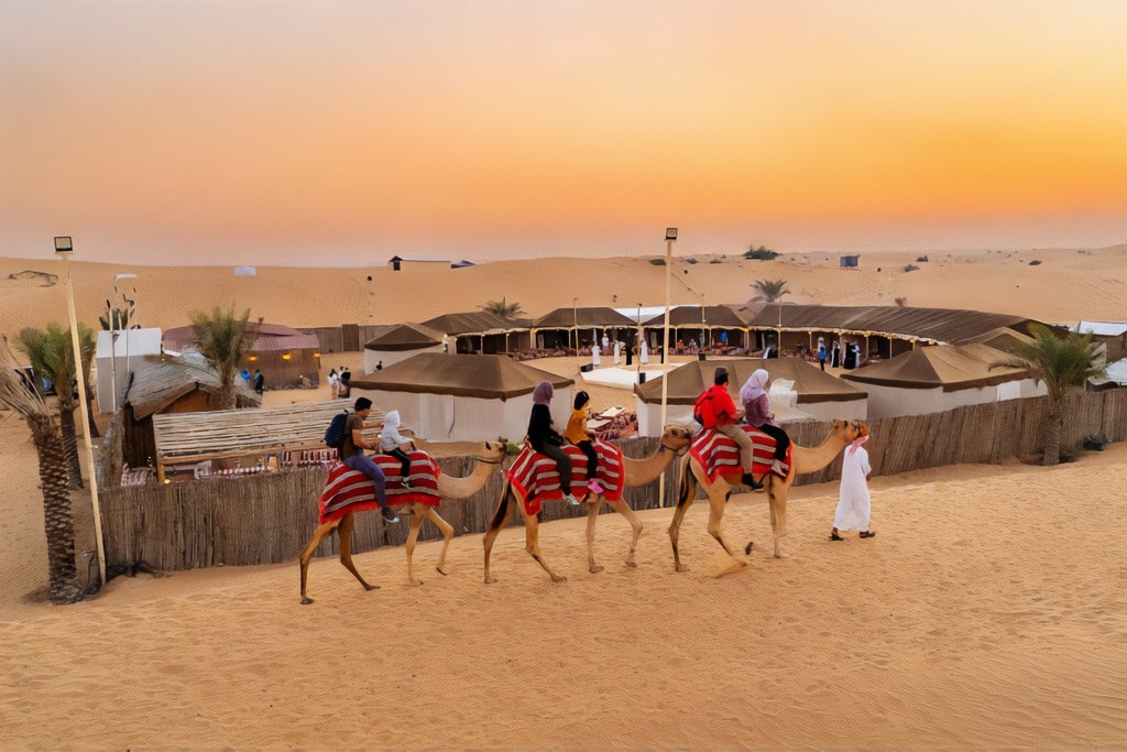 People riding camels in the desert near a traditional camp at sunset, with palm trees and tents in the background.