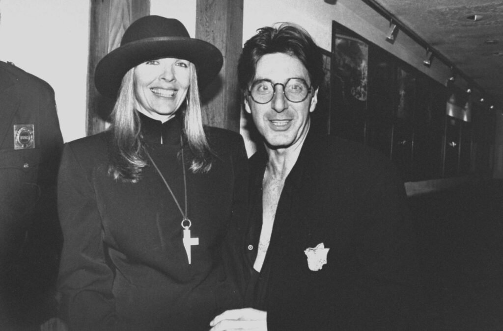 A black and white photo of Al Pacino and Diane Keaton smiling together. Diane Keaton is wearing a wide-brimmed hat and a necklace with a large cross pendant. Al Pacino is wearing glasses and a black shirt with an unbuttoned collar.