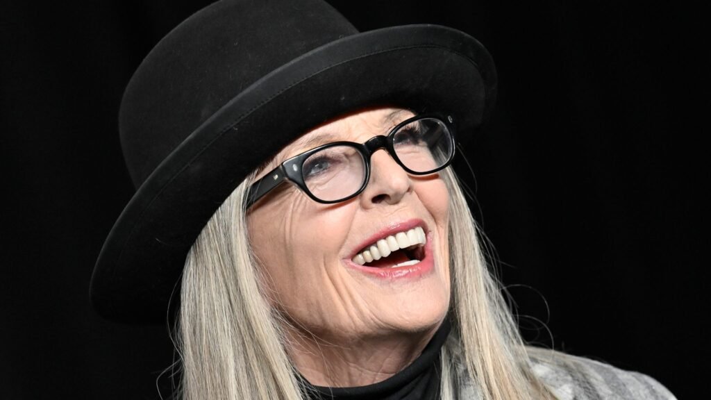 A person wearing a black hat and glasses, smiling broadly against a dark background.